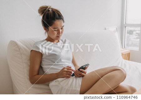 Woman in white t-shirt using mobile app sits on the sofa on a relax day in the morning. Woman in white t-shirt using mobile app sits on the sofa on a relax day in the morning. 110964746