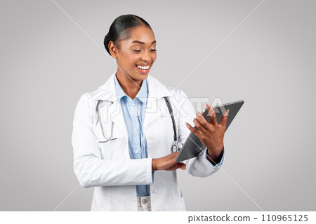 Smiling pretty millennial black woman doctor therapist in white coat typing on tablet, chatting 110965125