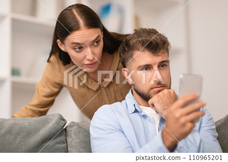 Jealous Girlfriend Spying On Boyfriend While Chatting On Smartphone Indoors 110965201