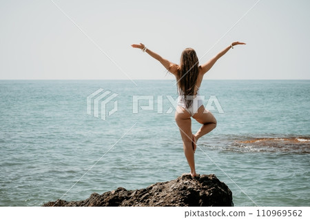 Woman sea yoga. Back view of free calm happy satisfied woman with long hair standing on top rock with yoga position against of sky by the sea. Healthy lifestyle outdoors in nature, fitness concept. Woman sea yoga. Back view of free calm happy satisfied woman with long hair standing on top rock with yoga position against of sky by the sea. Healthy lifestyle outdoors in nature, fitness concept. 110969562