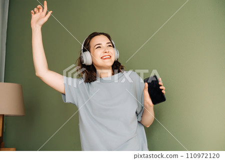 Happy young woman wearing wireless headphones listening to music, enjoying free time, dancing 110972120