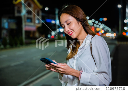 A woman stands on the sidewalk at night, holding her cellphone while waiting for transportation, with a vibrant orange and teal color scheme. A woman stands on the sidewalk at night, holding her cellphone while waiting for transportation, with a vibrant orange and teal color scheme. 110972718
