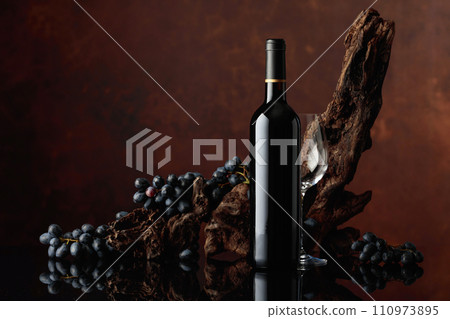 Bottle and glass of red wine with an old snag and blue grapes. 110973895