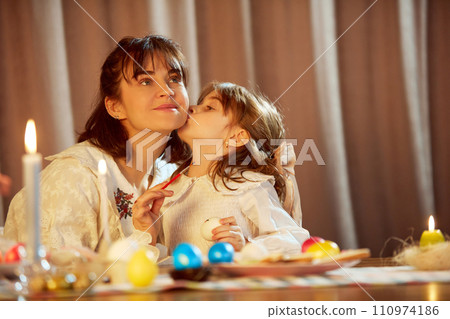 Little girl, child kissing her beautiful happy mother. Family sitting at table at home and painting Easter eggs. Coziness Little girl, child kissing her beautiful happy mother. Family sitting at table at home and painting Easter eggs. Coziness 110974186