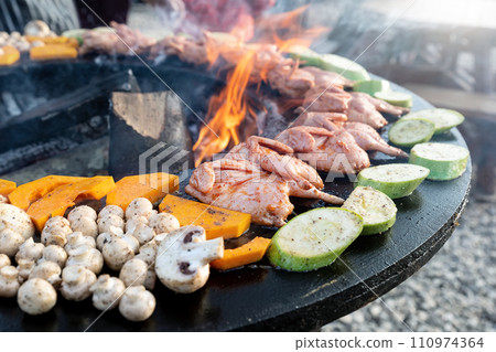 Close-up detail view of many tasty raw marinated quails and vegetables and mushrooms grilled cooked at round steel iron firepit hearth table surface brazier with burning firewoods. Barbecue yard home 110974364
