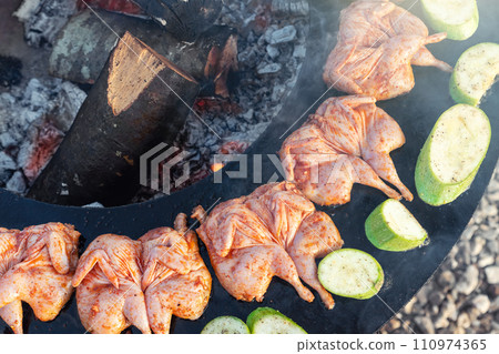 Top above view of many tasty raw marinated quails and vegetables and mushrooms grilled cooked at round steel iron firepit hearth table surface brazier with burning firewoods. Barbecue yard home 110974365