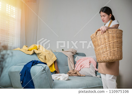 Asian housewife woman holding basket with heap of different clothes on sofa in living room, Beautiful housekeeper doing housework holds wooden basket of clean clothes to washing at home Asian housewife woman holding basket with heap of different clothes on sofa in living room, Beautiful housekeeper doing housework holds wooden basket of clean clothes to washing at home 110975106