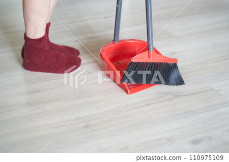 Side view of a red broom and plastic dustpan on the floor with person's legs in red socks. Cleaner, worker is using cleaning supplies with copy space. Concept of cleaning home, dust, sweeping floor. Side view of a red broom and plastic dustpan on the floor with person's legs in red socks. Cleaner, worker is using cleaning supplies with copy space. Concept of cleaning home, dust, sweeping floor. 110975109