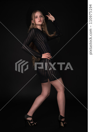 Blonde fashion model looks amazing in black short knitted translucent dress, wide-brimmed felt hat, high heels. Full length portrait captures her from three-quarter rear view against black background Blonde fashion model looks amazing in black short knitted translucent dress, wide-brimmed felt hat, high heels. Full length portrait captures her from three-quarter rear view against black background 110975194