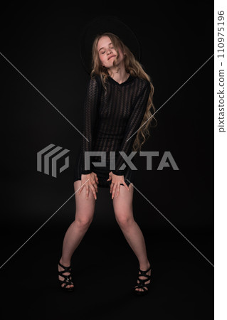 Confident young woman exuding elegance and style. With her eyes closed and lower lip pursed. She is wearing black short knitted translucent dress, wide-brimmed felt hat and high-heeled shoes Confident young woman exuding elegance and style. With her eyes closed and lower lip pursed. She is wearing black short knitted translucent dress, wide-brimmed felt hat and high-heeled shoes 110975196