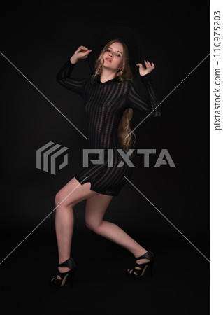 Fashion model with long legs. Full length portrait is set against black background. Sensuality blond with long hair is wearing black short transparent dress, felt hat with wide brim, high heeled shoes Fashion model with long legs. Full length portrait is set against black background. Sensuality blond with long hair is wearing black short transparent dress, felt hat with wide brim, high heeled shoes 110975203