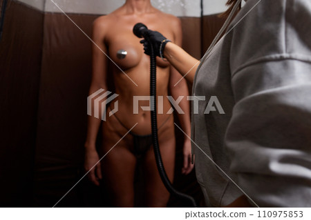 Cosmetologist using airbrush for spray tan apply to young woman body in beauty salon. 110975853