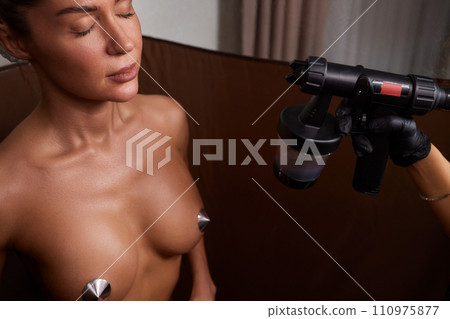 Cosmetologist using airbrush for spray tan apply to young woman body in beauty salon. 110975877