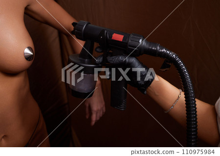 Cosmetologist using airbrush for spray tan apply to young woman body in beauty salon. 110975984