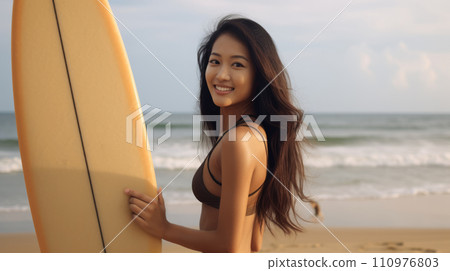 Happy Asian girl standing on the beach with a surfboard on a bright summer day. Smiling young Japanese woman posing on the shore with a board. Portrait of a cheerful Chinese surfer girl on a vacation. 110976803