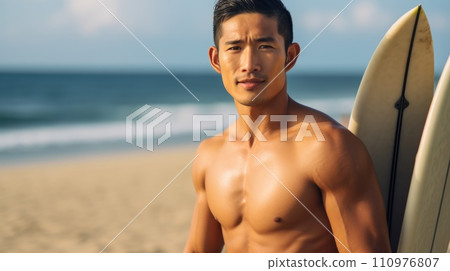 Happy Asian man standing on the beach with a surfboard on a bright summer day. Smiling muscular young Asian guy posing on the shore with a board. Portrait of a fit Chinese surfer on a vacation. 110976807
