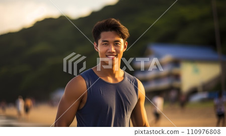 Happy Asian man standing on the beach on a bright summer day. Smiling muscular young Asian guy posing at the beach. Portrait of a fit Chinese surfer on a vacation. 110976808