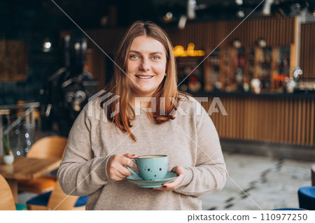 Attractive female in glasses with cute smile having a coffee while relax in in a break. Positive woman in casual wear smiling and resting with cup of hot drink in cozy cafe. Concept of easy breakfast Attractive female in glasses with cute smile having a coffee while relax in in a break. Positive woman in casual wear smiling and resting with cup of hot drink in cozy cafe. Concept of easy breakfast 110977250