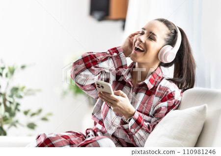 Smiling young woman sitting on couch with cell phone listening music with headphones Smiling young woman sitting on couch with cell phone listening music with headphones 110981246