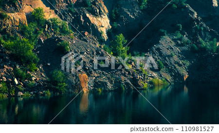 A rocky hillside with green shrubs near a clear, reflective water body. A rocky hillside with green shrubs near a clear, reflective water body. 110981527