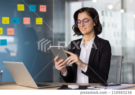 Businesswoman in business suit with tablet computer inside office at workplace, woman boss using app, sitting at table with laptop, happy with work smiling. 110983236