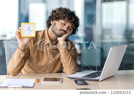 Exhausted muslim man sitting at laptop with closed eyes and showing alarm clock in one hand. Young male in shirt sleeping at office desk during break and holding timer for not be late to meeting. Exhausted muslim man sitting at laptop with closed eyes and showing alarm clock in one hand. Young male in shirt sleeping at office desk during break and holding timer for not be late to meeting. 110983253