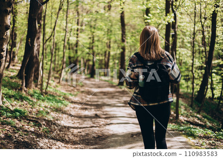 Rear view of young woman with casual outfit, wears a black backpack walking in the forest. Spring time. 30s Women in sport clothes hiking in forest, back view. Wanderlust travel concept 110983583