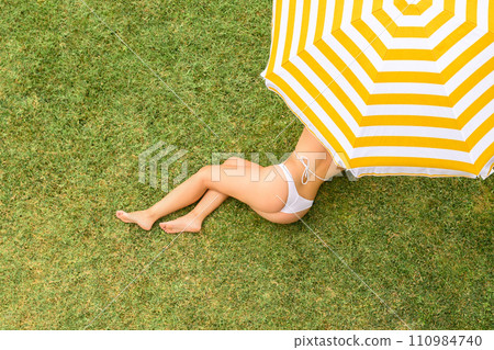Woman in white bikini lying under yellow umbrella on the green grass sunbathes at summer day. Top view, drone, aerial view. 110984740
