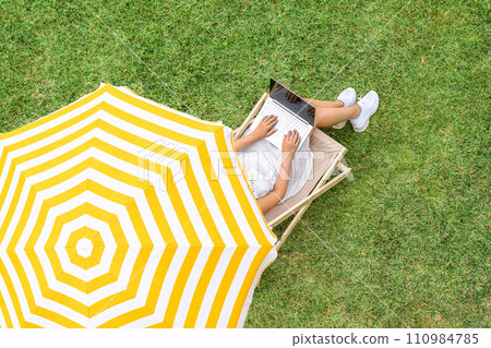 Woman in a white dress sitting on deck chair under yellow umbrella with laptop on the green grass sunbathes at summer day. Top view, drone, aerial view. 110984785