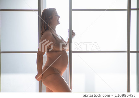 A pregnant woman in nude lingerie stands in profile between folding doors. 110986455