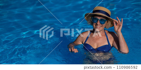 Portrait of an elderly woman in a hat and blue sunglasses swims in the pool. Portrait of an elderly woman in a hat and blue sunglasses swims in the pool. 110986592