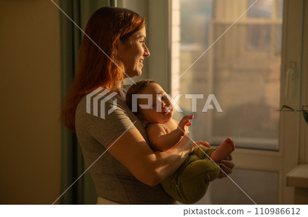 Caucasian woman tenderly holds her newborn son while standing near the window. Caucasian woman tenderly holds her newborn son while standing near the window. 110986612