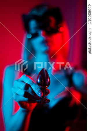 Blonde woman wearing a leather mask in red blue neon light and holding a steel butt plug.  110986649