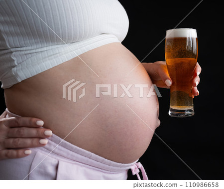 Close-up of the belly of a pregnant woman holding a glass of beer on a black background. 110986658