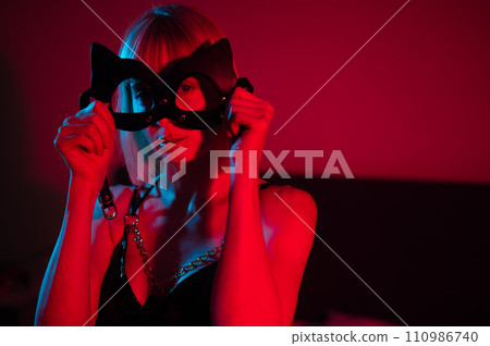 Blonde woman putting on leather mask in bedroom in red blue neon light. Blonde woman putting on leather mask in bedroom in red blue neon light. 110986740