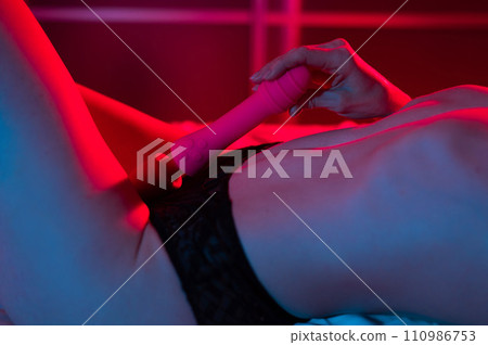 A faceless woman lies in a bedroom in a blue-red neon light and uses a pink vibrator. Close-up of the abdomen. A faceless woman lies in a bedroom in a blue-red neon light and uses a pink vibrator. Close-up of the abdomen. 110986753