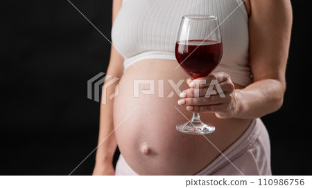 Close-up of the belly of a pregnant woman holding a glass of red wine on a black background.  110986756