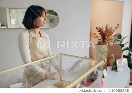 Beautiful female barista is looking away and smiling while standing near bar counter in cafe 110988208