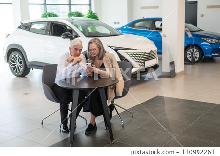 A mature couple is sitting in a car dealership and looking at a smartphone.  110992261