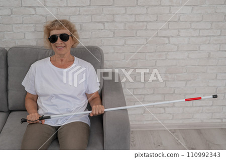 An elderly blind woman wearing sunglasses and with a tactile cane sits on the sofa.  110992343