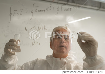 An elderly Caucasian male chemist in a protective suit writes on glass.  110992370