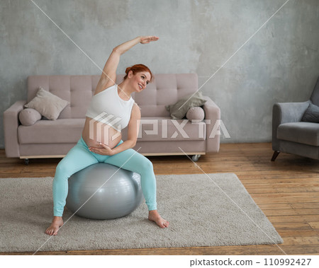 Pregnant red-haired woman doing arm exercises with fitball at home. 110992427
