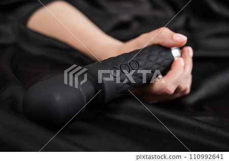 A woman is holding a black vibrator while lying on a black silk sheet. A woman is holding a black vibrator while lying on a black silk sheet. 110992641