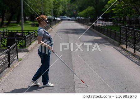 Blind pregnant woman crosses the street with the help of a tactile cane. 110992670