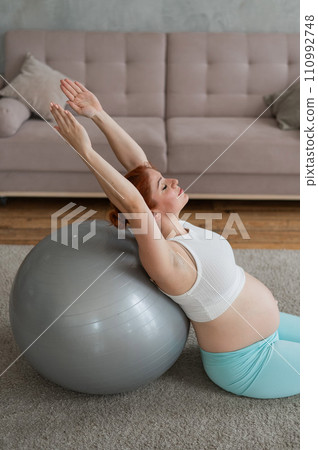 Pregnant red-haired woman doing exercises on fitness ball at home.  110992748