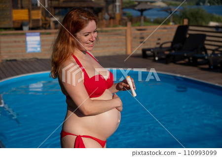 A red-haired pregnant woman sunbathes by the pool. The expectant mother applies sunscreen to her tummy. A red-haired pregnant woman sunbathes by the pool. The expectant mother applies sunscreen to her tummy. 110993099