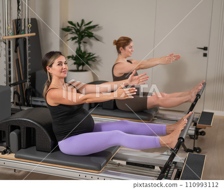 Two pregnant women do Pilates exercises on a reformer. Yoga class for pregnant women. Two pregnant women do Pilates exercises on a reformer. Yoga class for pregnant women. 110993112