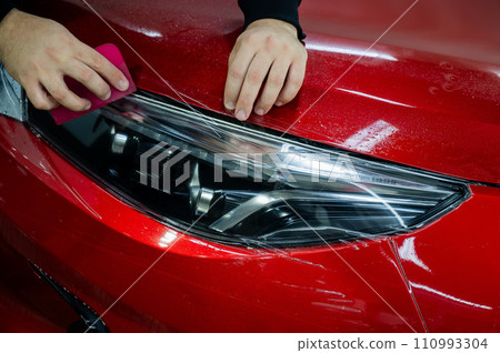 The master applies vinyl film to the headlight of a red car. Closeup view on worker detailer hand smoothing with a scraper protective film.  110993304