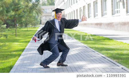 Old happy man in graduation gown jumping outdoors and holding diploma. 110993391