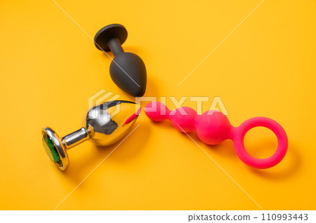 A set of three alternative sex devices of various shapes and sizes on an orange background. Silicone and metal plugs and anal beads. A set of three alternative sex devices of various shapes and sizes on an orange background. Silicone and metal plugs and anal beads. 110993443